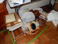 Photo showing baskets containing needlework supplies including knitting needles, fabric projects, and sewing items alongside stacked picture frames and plastic bags.
