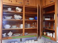 Wooden kitchen cabinet shelves containing an assortment of dishware: white porcelain bowls, hand-painted floral serving pieces, various glassware, and blue decorative bowls in top right corner.