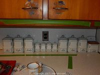 Photo showing full canister set with 8 large canisters, 4 small spice canisters, and 1 salt box arranged on kitchen counter against wood cabinets.