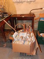 Full view of the wooden sewing machine table and the cardboard box filled with sewing patterns and notions nearby on the floor in a basement.