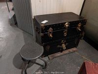 Two black travel trunks stacked vertically with brass locks and studs. A metal stool with perforated round seat is positioned to the left of the trunks. Setting appears to be in basement with concrete floor and white paneling on wall.