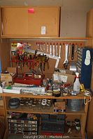 Wide shot of wooden workstation with hand tools hanging on pegboard, metal hardware drawers, mason jars, and miscellaneous items on the tabletop, shelves below contain more tools and hardware.