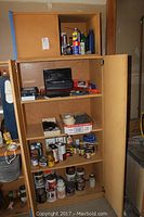 Wooden cabinet with multiple shelves containing woodworking chemicals, wood finishes, wood stains, cans, bottles, and other supplies.