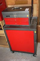 Metal toolbox sitting on top of rolling tool storage caddy, showing red and gray paint, toolbox handle, and latch.
