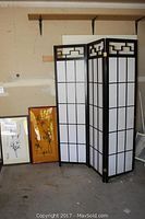 Three-panel black wooden Shoji screen standing with two framed Asian ink drawings leaning against the wall, visible wear and dust.