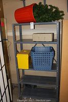Metal shelving unit with four gray metal shelves, containing plastic baskets, yellow filing box, beige storage box, and artificial tree in red container on top shelf.