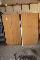 Two light brown wooden storage cabinets standing side by side with closed double doors in a garage setting.