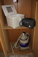 Air compressor cooler, AC 120 Volt adapter, and rolling shop vac stored on wooden shelves. Cooler is white with lid open. Shop vac is white and blue with hose attached.