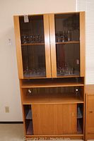 Front view of the modern maple colored cabinet showing glass doors with glassware inside, open shelf, and sliding wooden doors at the bottom.