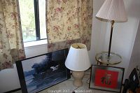 Photo showing mid century round glass lamp table with brass accents and lamp with white pleated shade, beige ceramic table lamp, two framed art pieces with one featuring red Chinese character, and framed landscape print of duck on lake