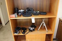 Wide view of bookshelf shelf containing cameras, tripod, electronic flash, camera cases, lenses, and light meter