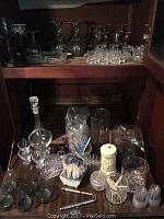Two wooden shelves displaying an assortment of glassware and barware including stemmed glasses, short glasses, decanter, various small bowls, and bar accessories.