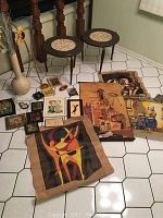 Two round wooden end tables with three legs, dark wood top with circular inlay, brass accent rings visible on legs, right near a large beige vase with dried flowers and various decorative framed and unframed textile art pieces arranged on floor.