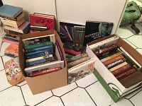 Photo showing several boxes and stacks of books on the floor, some hardcovers about astronomy, chemistry, and classics.