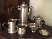 Photo showing the full set of five pewter items on a dark surface: coffee pot, sugar bowl, creamer, tankard, and plate.