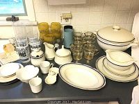 Full collection of Malowane china dinnerware pieces displayed on a counter including plates, casserole, gravy boat, sugar bowl, creamers, and glassware of yellow and clear glasses.