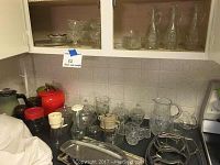 Full view of glassware on kitchen counter under cabinets showing multiple pitchers, candy dishes, glasses, and trays.