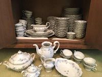 Full view of the china set including stacked plates, cups with saucers, teapot, sugar bowl, creamer, and covered tureen in white porcelain with light blue floral pattern and gold trim.