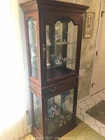 Full view of the tall wooden china cabinet with glass doors, two glass shelves in upper and lower compartments, and central wooden drawer with brass handles.