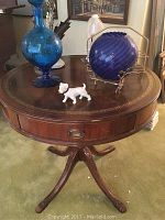 Front view of round wood end table holding blue crackle glass bottle, cobalt blue glass orb on metal stand, and white porcelain dog figurine