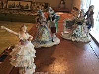 Photograph of three German-made porcelain figurines displayed on a wooden table, two couples in 18th-century style dress and one ballerina figurine.