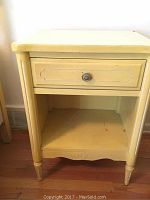Front view of vintage yellow end table displaying single drawer with decorative knob and open lower shelf. Shows some wear and paint marks on lower shelf and edges.