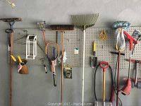 Gardening tools hanging on pegboard including shovels, rakes, small hand tools, tennis racket, and two red rolling carts.