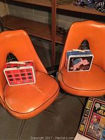 Two vintage orange chairs with two retro metal lunchboxes placed on them, one with a red plaid pattern and the other with an American flag and eagle design.