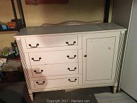 White wooden dresser in basement with four drawers on the left and one cabinet door on the right. Visible wear marks on top surface and scuffing on legs.