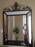 Front view of large beveled mirror showing reflective surface, distressed metal frame, and decorative top scroll design with fleur-de-lis accent.