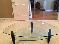 Front angle view of geometric two-tier glass coffee table with black tapered legs on beige carpet in room with white door and hardwood floor in background.