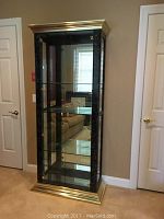 Lighted glass display cabinet with gold base, crown molding, and faux marble pattern. Shows front side with multiple glass shelves inside, glass door with gold knobs, and visible interior light.