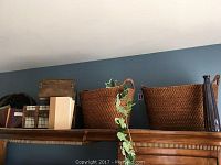 Old world style decor items on a wooden cabinet against a blue wall featuring 2 wicker baskets, 2 wooden trunks with plaid sides, and several books.