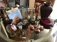 Full view of kitchen decor items arranged on a round glass-top table including ceramic bottles, oil and vinegar bottles, chef figurines, a red and black ceramic dish with lid, a decorative clock with grape motif, two candle holders, and cookbooks.