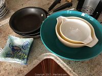 Photo showing two copper bottom frying pans, one large teal Pyrex bowl containing two smaller bowls (white and yellow), and stack of 6 patterned glass square dishes with floral and peacock designs on countertop.