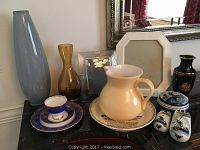 Photograph shows a collection of decor items on a black surface including a tall blue Limoges vase, an amber glass vase, a cream-colored pitcher, Delft pottery including salt and pepper containers, a blue and gold tea set, a white octagonal frame, and a small black vase with gold floral design.