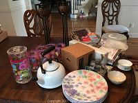 Wide shot of various kitchenware items including Le Creuset tea kettle, floral dinner plates, glass pitcher, juice cups, electric knife box, and additional decor on wooden table.