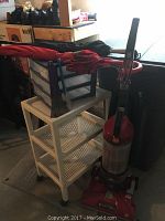Photo of red Dirt Devil upright vacuum cleaner, white rolling cart with mesh shelves, blue plastic storage bins with drawers, and a red umbrella resting on top of the cart