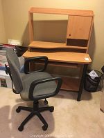 Front view of the desk and swivel chair showing the desk's light wood finish, upper hutch with cabinet, and black frame. Chair is gray upholstered with armrests and caster wheels.