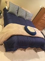 Full size bed made up with navy blue comforter, beige bed skirt, three decorative pillows in black and white pattern, and matching beige throw blanket with round pillow on top of bed.