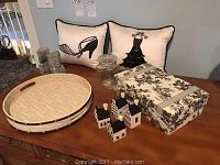 Photo showing two decorative pillows with black designs (shoe and dress), a glass jar, three ceramic house-shaped bottles, a large rectangular fabric storage box with floral toile pattern, and a round wooden tray on a wooden surface.