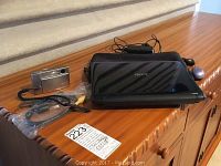 Acer Aspire One mini laptop in black with charger and case, Sony Cybershot digital camera, and cables on a wooden dresser.