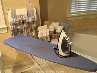 Full view of ironing board with blue cover and iron placed on top, baskets of towels visible in background on floor