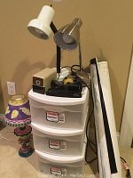 Full view of plastic storage drawers with three desk lamps and various office supplies on top, including stapler and tape dispenser. Decorative lamp with purple-yellow shade is on the side.