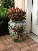 Plant ceramic planter positioned on tiled floor with pink flowers planted inside. Signed hand painted colorful Asian figures, birds, and flowers on blue background.