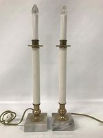 Full view of the two electric lamps showing white candle-like columns with flame-shaped bulbs on marble bases with brass holders.