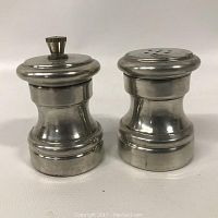 Photo showing front view of two pewter shakers, one with perforated lid and one with removable knob lid.