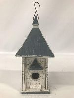 Front view of the wooden birdhouse showing the triangular blue roof, circular entry hole, small platform, and crackled white paint on the body.