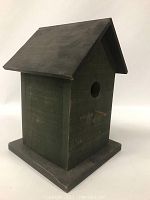 Side angled view showing dark forest green wooden bird house with peaked dark roof and base, circular entrance hole, and small wooden perch below the hole.