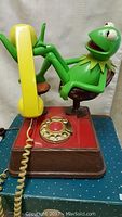 Side view of Kermit the Frog figure reclining on chair on phone base with yellow handset visible.